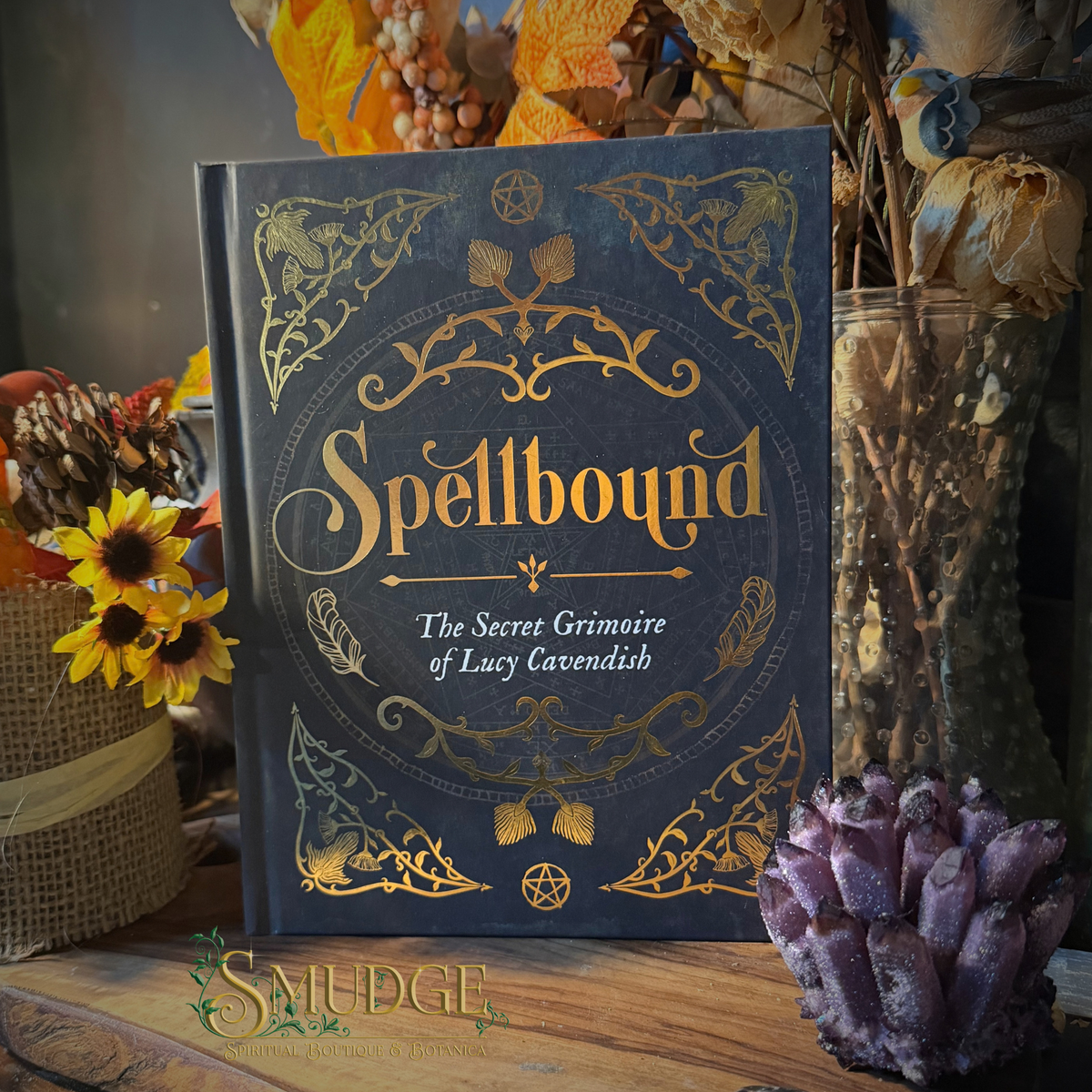 A hardcover book titled "Spellbound: The Secret Grimoire of Lucy Cavendish" stands upright on a wooden surface, surrounded by autumn-themed decorative props including dried flowers, sunflowers, and a purple crystal cluster. The cover is dark blue with ornate gold illustration and lettering.