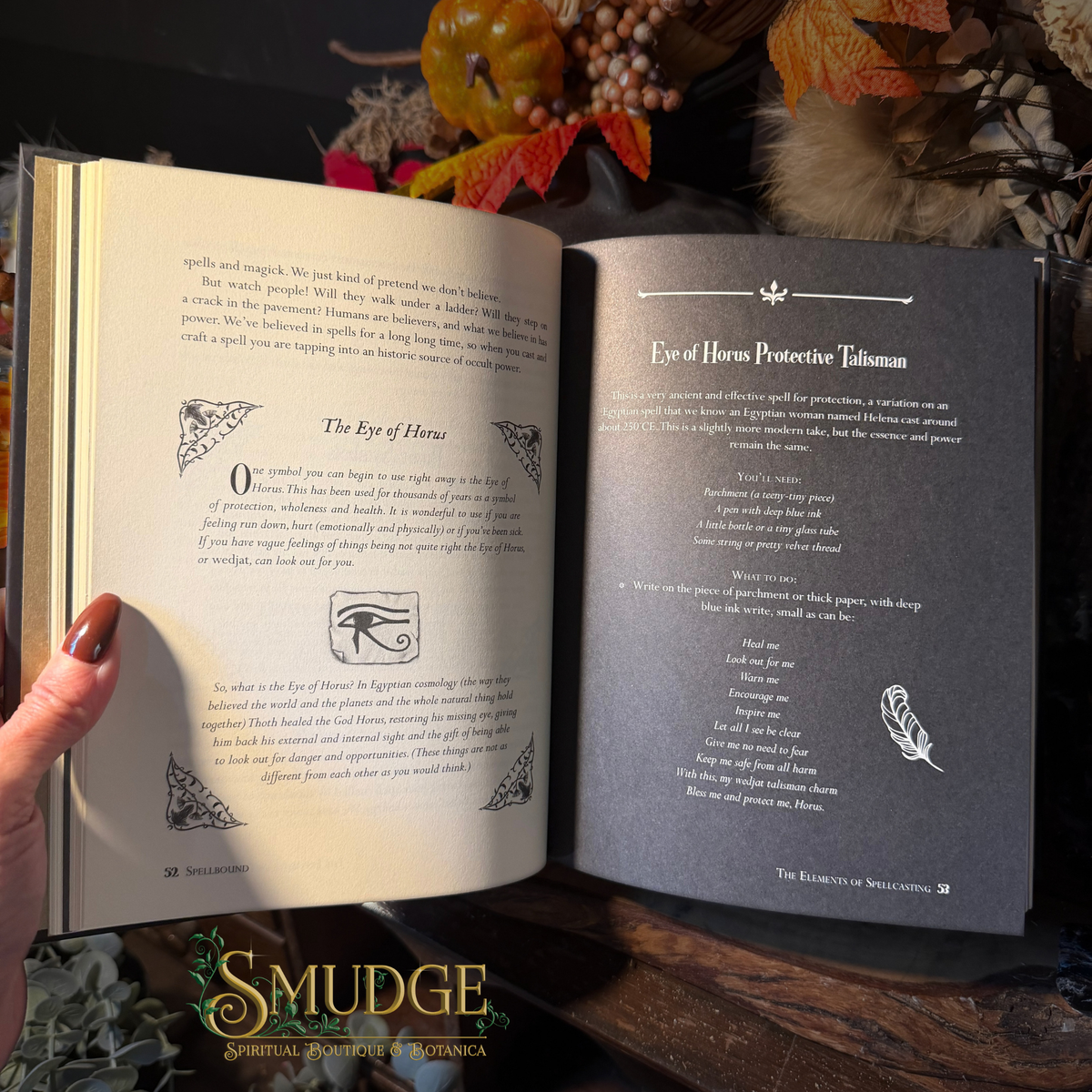 An open book showing two printed pages with text and decorative illustrations, held by a hand with dark red nail polish. The setting is a styled tabletop with autumn leaves, dried flowers, and a wooden surface.