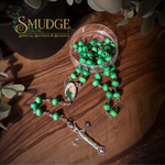 A green beaded rosary with a silver-tone crucifix and a central religious medallion, arranged on a wooden surface next to a small clear container of beads. The brand name 'SMUDGE' is visible in the upper left corner.