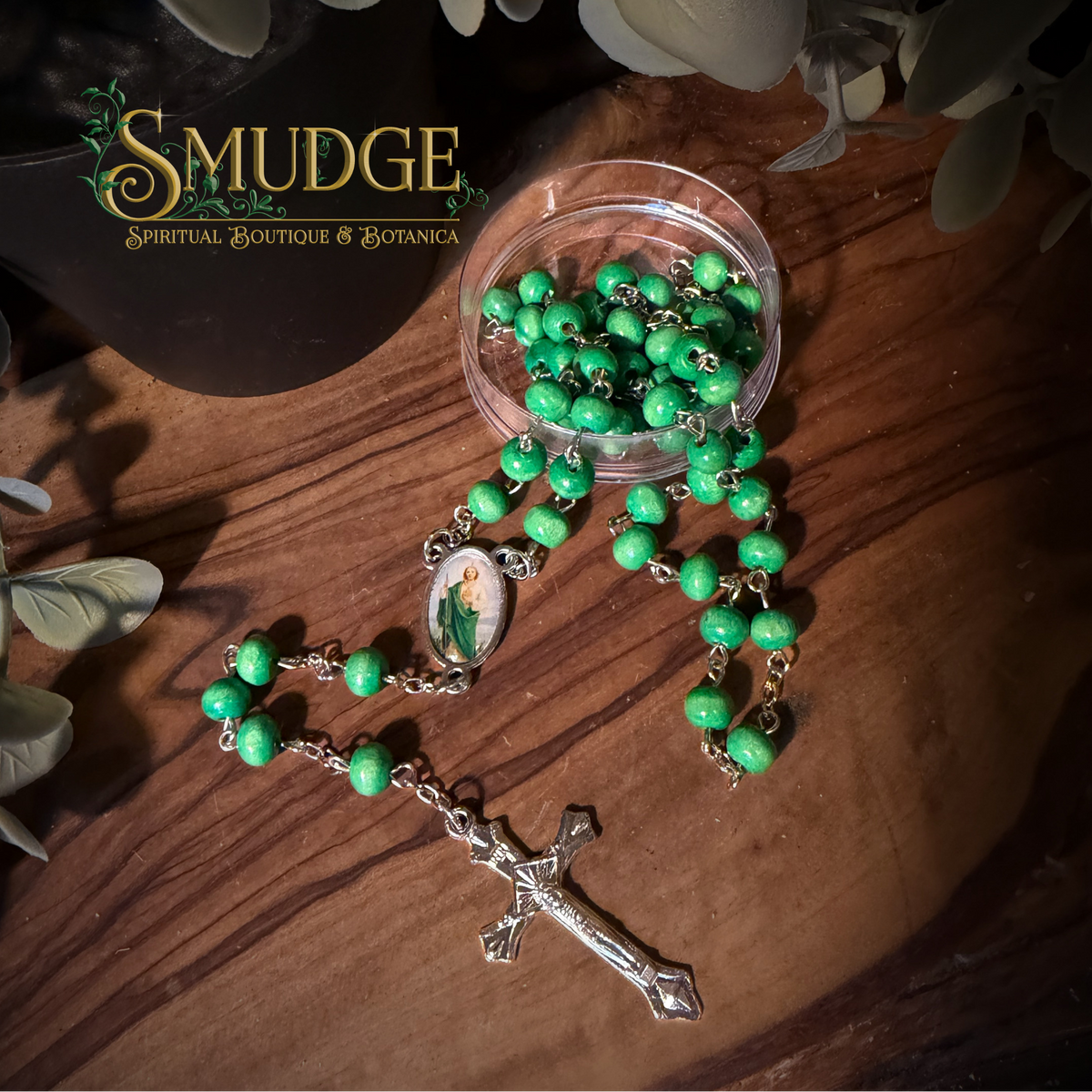 A green beaded rosary with a silver-tone crucifix and a central religious medallion, arranged on a wooden surface next to a small clear container of beads. The brand name 'SMUDGE' is visible in the upper left corner.
