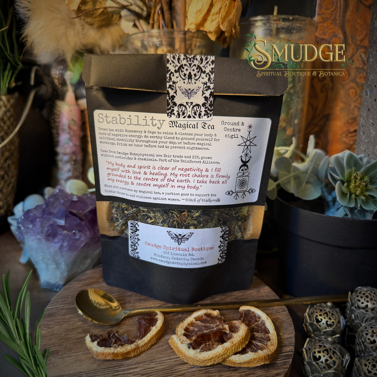 A black stand-up pouch of Stability Magick Tea by Smudge Spiritual Boutique is displayed on a wooden board with dried orange slices and a gold spoon. The background is a styled bohemian setting with crystals, dried flowers, and candles.