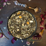 A top-down view of a black bowl filled with a loose herbal tea blend, surrounded by scattered dried flower petals on a dark wooden surface. An ornate shell-shaped spoon rests at the top, and the brand name 'MUDGE' is printed at the bottom right.