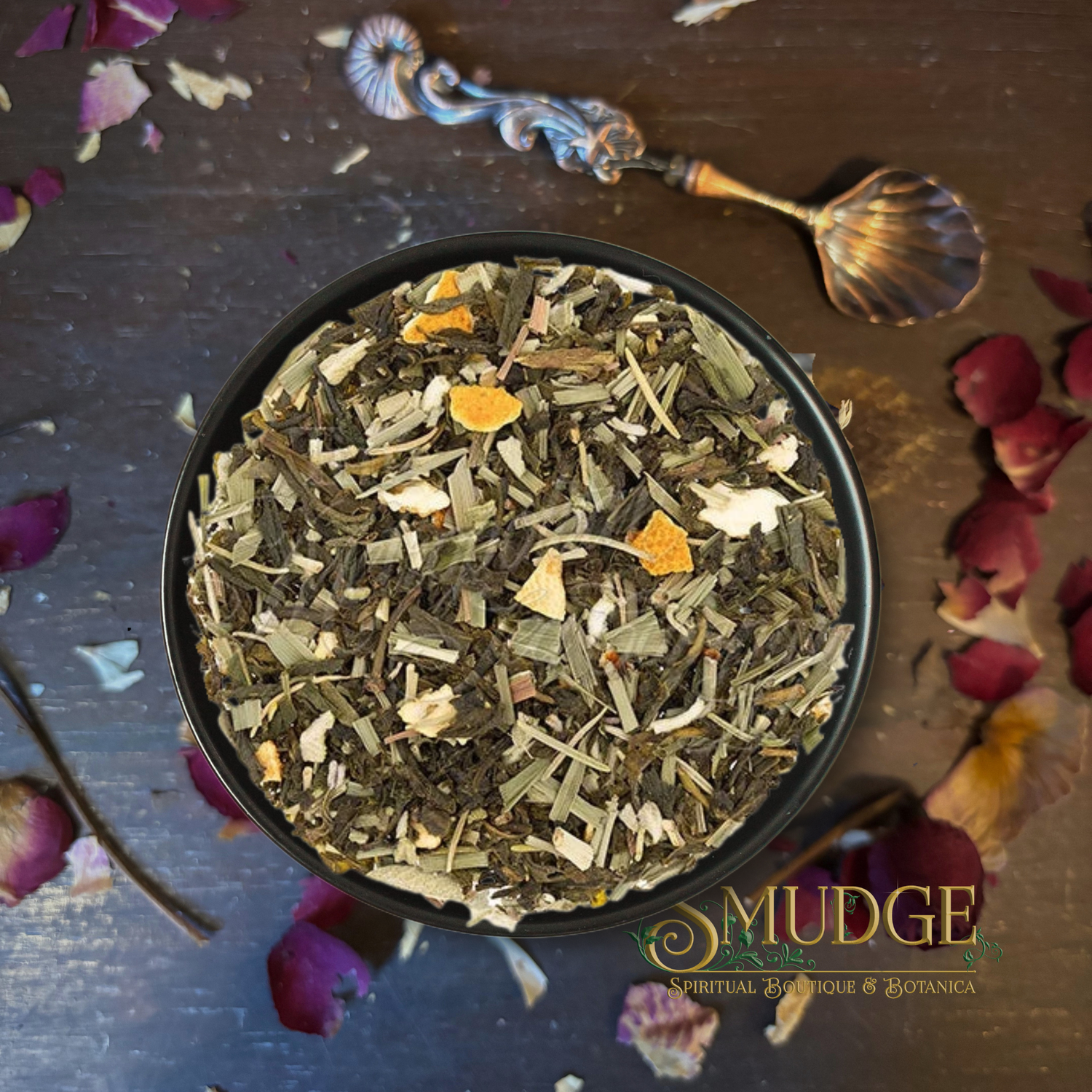 A top-down view of a black bowl filled with a loose herbal tea blend, surrounded by scattered dried flower petals on a dark wooden surface. An ornate shell-shaped spoon rests at the top, and the brand name 'MUDGE' is printed at the bottom right.