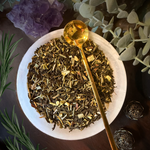 A top-down view of a white ceramic bowl filled with loose herbal tea blend, with a shiny gold spoon resting inside. The scene is styled with botanical elements including rosemary, eucalyptus, and an amethyst crystal cluster on a dark wooden surface.
