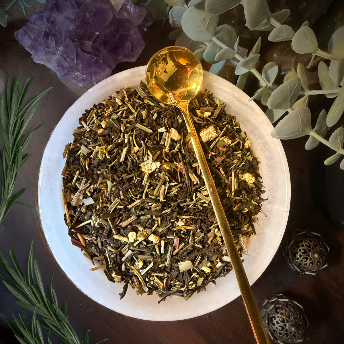 A top-down view of a white ceramic bowl filled with loose herbal tea blend, with a shiny gold spoon resting inside. The scene is styled with botanical elements including rosemary, eucalyptus, and an amethyst crystal cluster on a dark wooden surface.