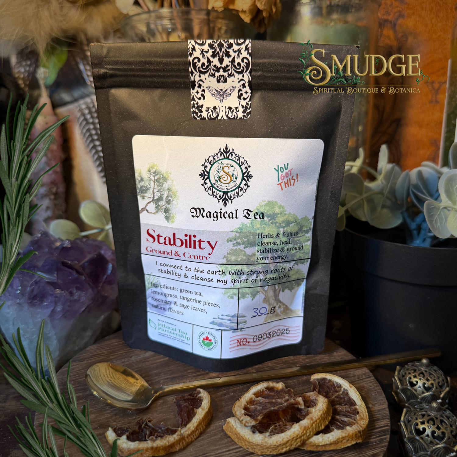 A stand-up pouch of Smudge Natural Herbs & Botany 'Stability' Ground & Coated Magical Tea, styled on a wooden surface with dried orange slices, crystals, and rosemary.
