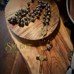 A dark brown and gold beaded rosary with a gold medallion and cross, displayed on a rustic wooden board. The brand logo 'Smudge Spiritual Boutique & Botanical' is visible on the lower left.