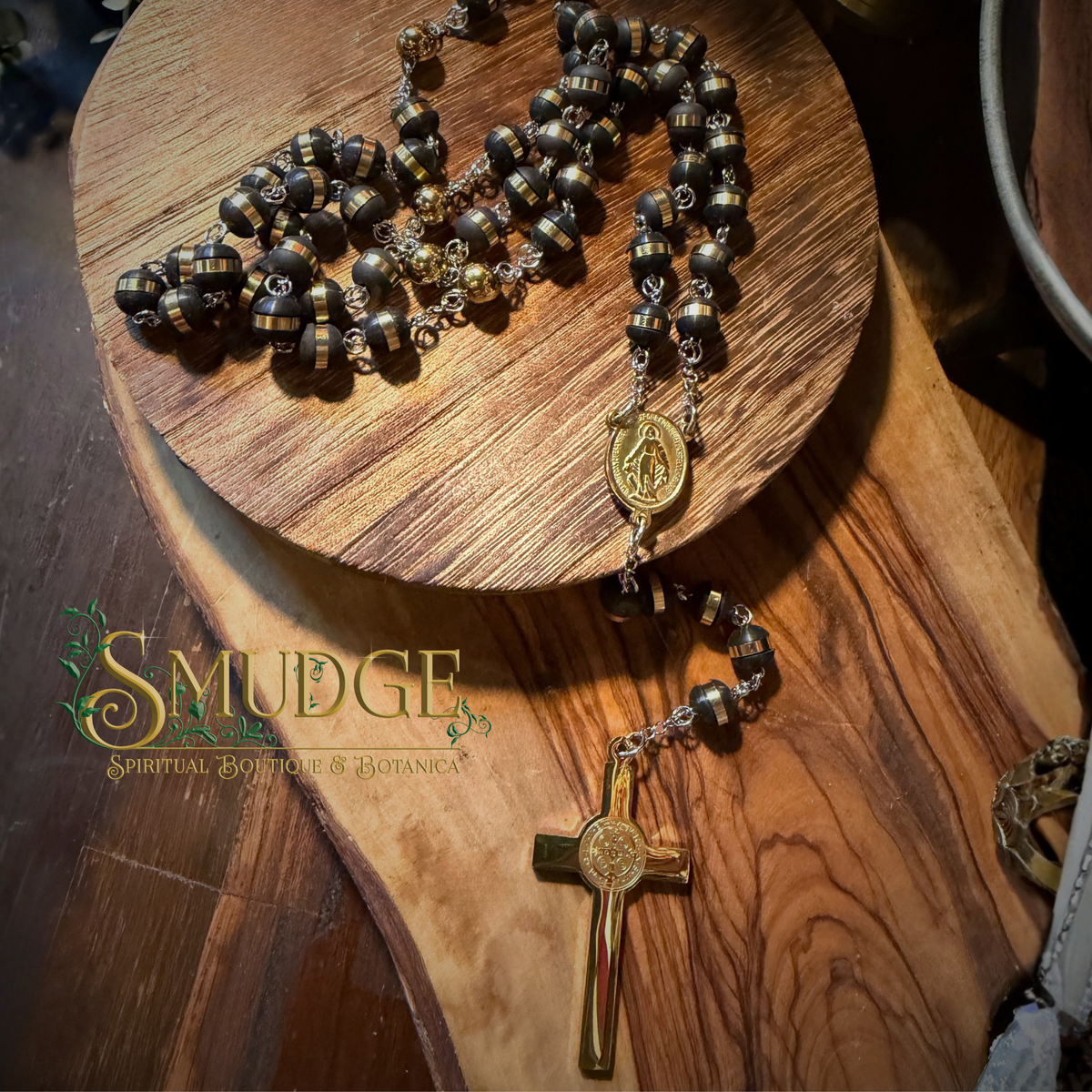 A dark brown and gold beaded rosary with a gold medallion and cross, displayed on a rustic wooden board. The brand logo 'Smudge Spiritual Boutique & Botanical' is visible on the lower left.