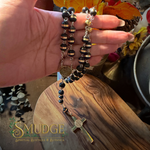 A hand holds a beaded rosary with black and metallic beads, a gold-toned cross, and a round medallion over a wooden surface. The words 'SMUDGE Spiritual Boutique & Botanica' appear as a watermark at the bottom left.