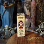 A small clear glass bottle of Bendictum Perfume labeled 'St. Clara' stands on a wooden surface surrounded by decorative props including crystals, feathers, dried leaves, and figurines in a styled indoor setting.