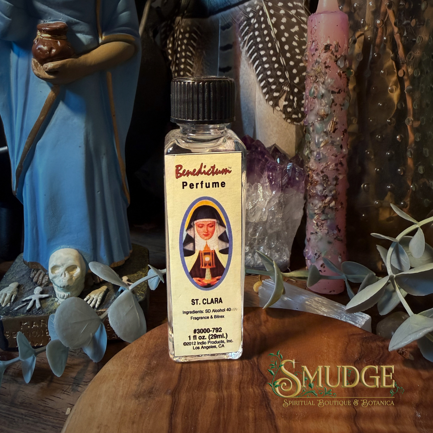 A small clear glass bottle of Bendictum Perfume labeled 'St. Clara' stands on a wooden surface surrounded by decorative props including crystals, feathers, dried leaves, and figurines in a styled indoor setting.