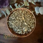 A copper-colored bowl filled with dried nettle leaf botanical herbs and small wood chips, photographed from above on a dark wooden surface with decorative leaves and crystals around it. The brand name 'SMUDGE' is visible at the bottom left.