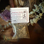 A clear plastic bag containing dried Stinging Nettle Leaf herbal powder, with a white paper label reading 'Magical Herbs' and 'Stinging Nettle Leaf Urtnella dioica.' The bag is photographed on a dark wooden surface with decorative plants, crystals, and a glass container in the background.