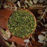 A top-down view of a brown ceramic bowl filled with finely chopped green herbal leaves, surrounded by dried flowers, greenery, and crystals on a dark wooden surface. The brand logo 'The Squeeze Botanicals' is visible in the top right corner.
