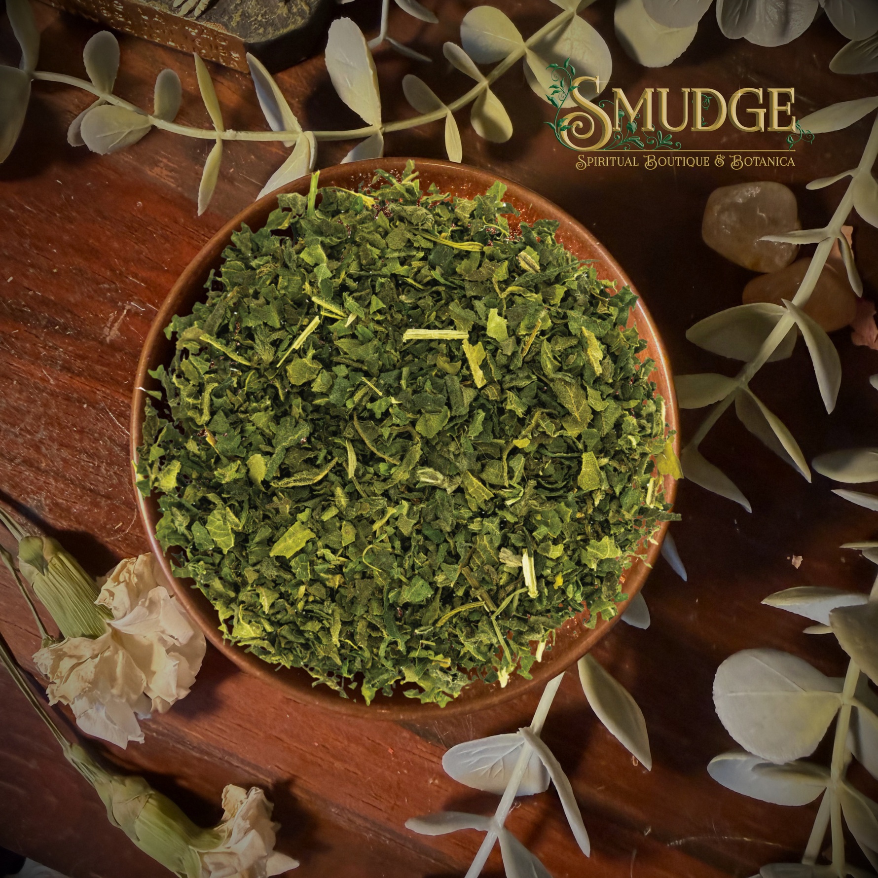 A top-down view of a brown ceramic bowl filled with finely chopped green herbal leaves, surrounded by dried flowers, greenery, and crystals on a dark wooden surface. The brand logo 'The Squeeze Botanicals' is visible in the top right corner.