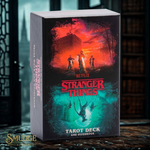 Stranger Things tarot deck and guidebook box set on a dark wooden table with a blurred library background. The box features Stranger Things artwork with red and teal tones, and the Smudge brand logo is visible in the bottom-left corner.