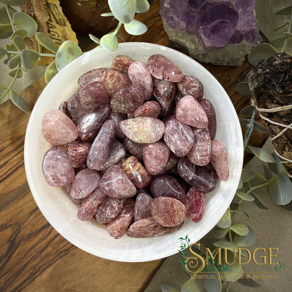 A white ceramic bowl filled with polished tumbled gemstones in pink, mauve, and reddish tones, styled on a wooden surface with green leaves and crystal clusters around it. A 'SMUDGE Spiritual Minerals & Botanica' logo is visible in the lower right corner.