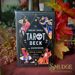 The Sugar Skull Tarot Deck and Guidebook box standing upright on a draped fabric surface, surrounded by autumn leaves and flowers. The box is black with colorful illustrated tarot card characters and white title text.