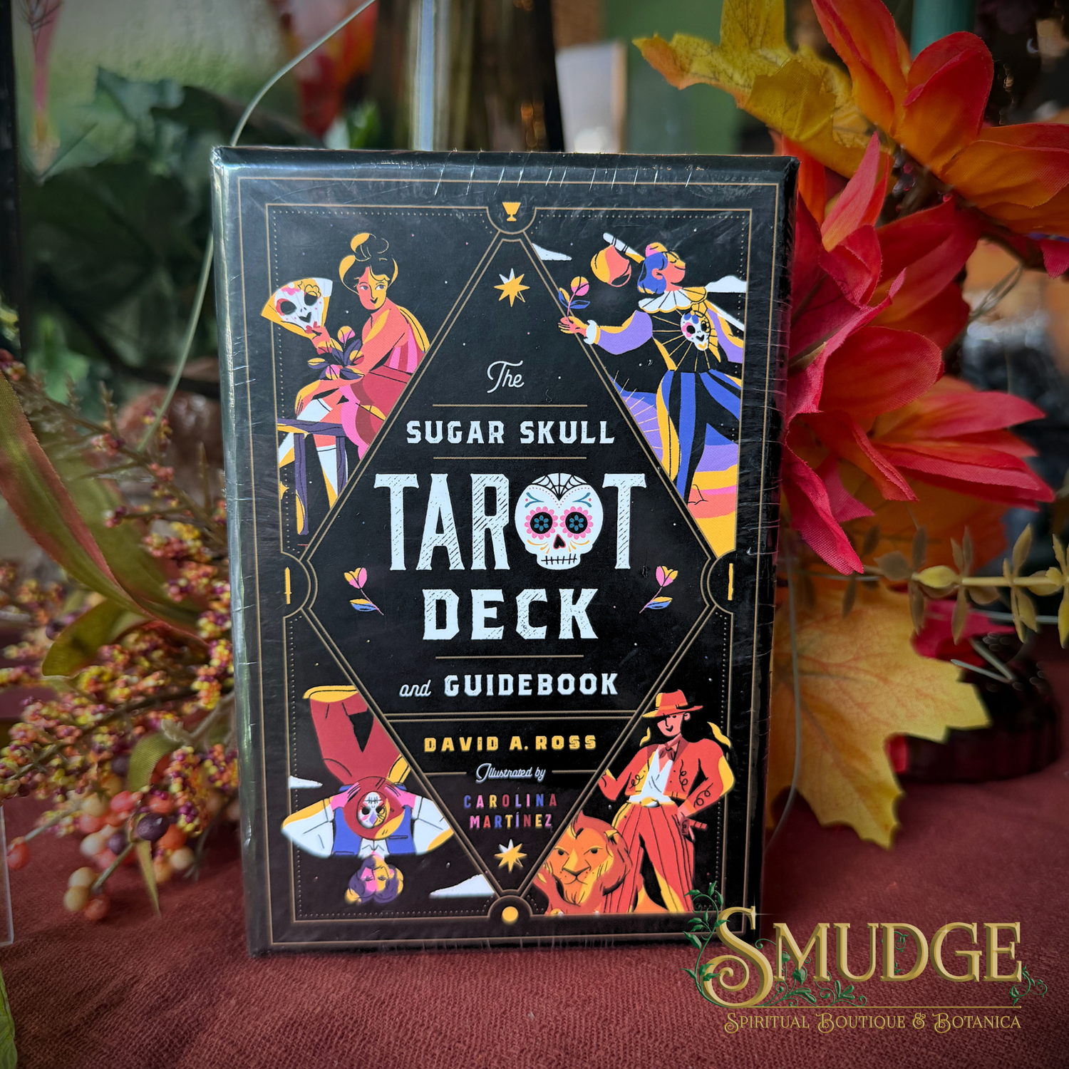 The Sugar Skull Tarot Deck and Guidebook box standing upright on a draped fabric surface, surrounded by autumn leaves and flowers. The box is black with colorful illustrated tarot card characters and white title text.