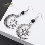 A pair of silver-toned crescent moon and sun dangle earrings with ornate filigree detailing, black beads, and silver fishhook wires, displayed on a white surface with a dark corner background. The brand logo 'SMUDGE Spiritual Jewelry & Botanica' is visible in the top left corner.