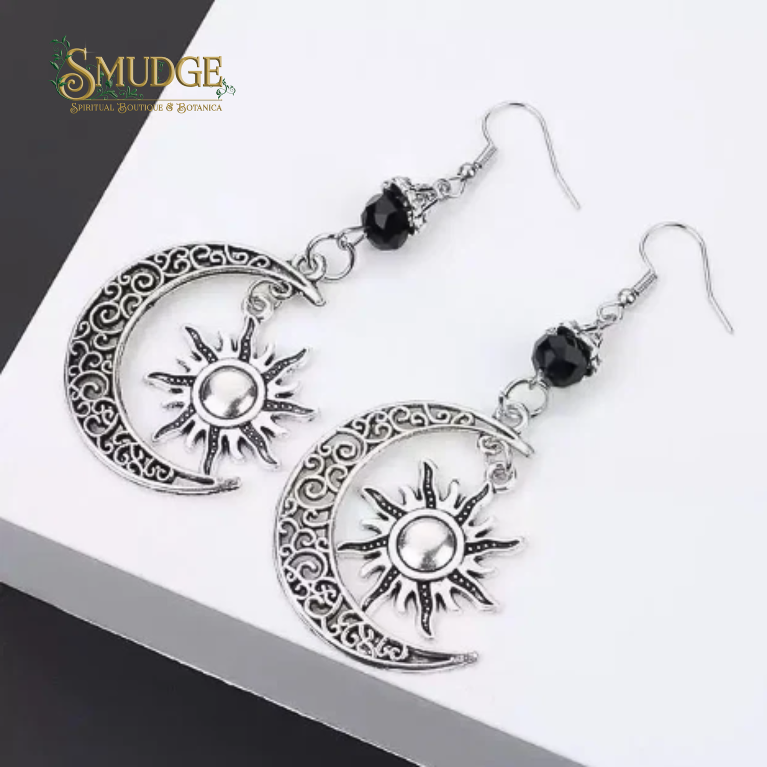 A pair of silver-toned crescent moon and sun dangle earrings with ornate filigree detailing, black beads, and silver fishhook wires, displayed on a white surface with a dark corner background. The brand logo 'SMUDGE Spiritual Jewelry & Botanica' is visible in the top left corner.