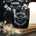 Black mug with 'Support Your Local Witch Coven' text on a wooden coaster.