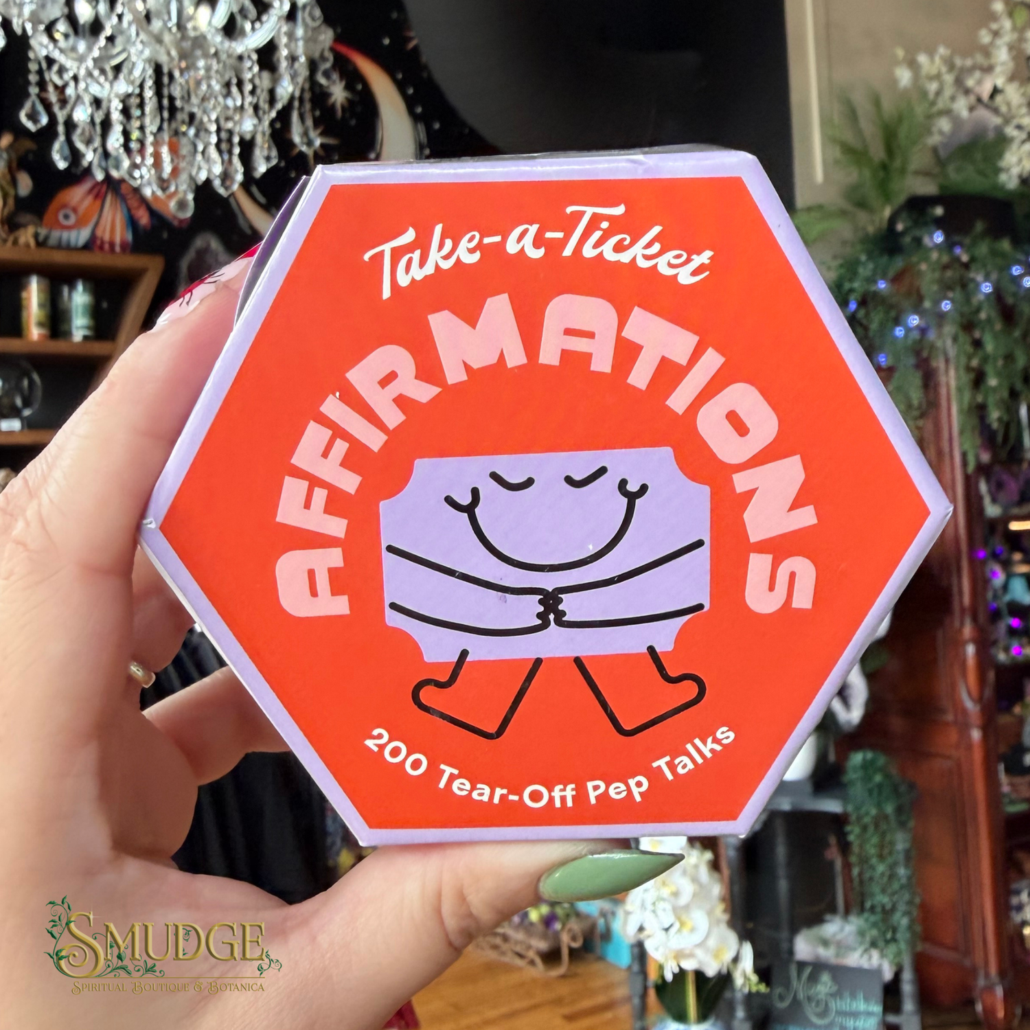 A hand holding a bright orange hexagonal product box labeled 'Take-a-Ticket AFFIRMATIONS' with a cartoon tear-off ticket graphic, in a blurred indoor boutique setting.