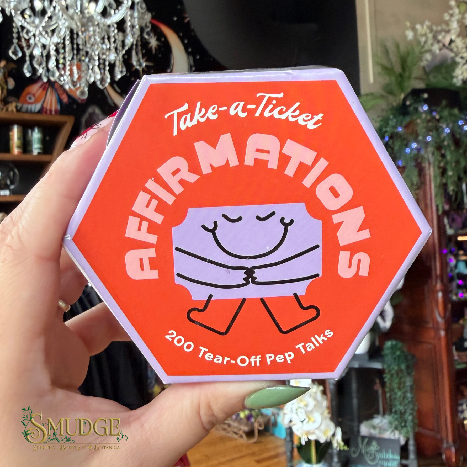 A hand holding a bright orange hexagonal product box labeled 'Take-a-Ticket AFFIRMATIONS' with a cartoon tear-off ticket graphic, in a blurred indoor boutique setting.