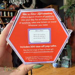 A hand holds a hexagonal orange product box for Take-a-Ticket affirmations, showing the back side with text and a small tear-off ticket sample. The background is a real indoor setting with shelves and decor visible.