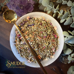 A top-down view of a white ceramic bowl filled with loose herbal tea or botanical blend, with a gold spoon resting across the bowl. The scene includes green leaves, purple crystals, and a dark wooden surface.