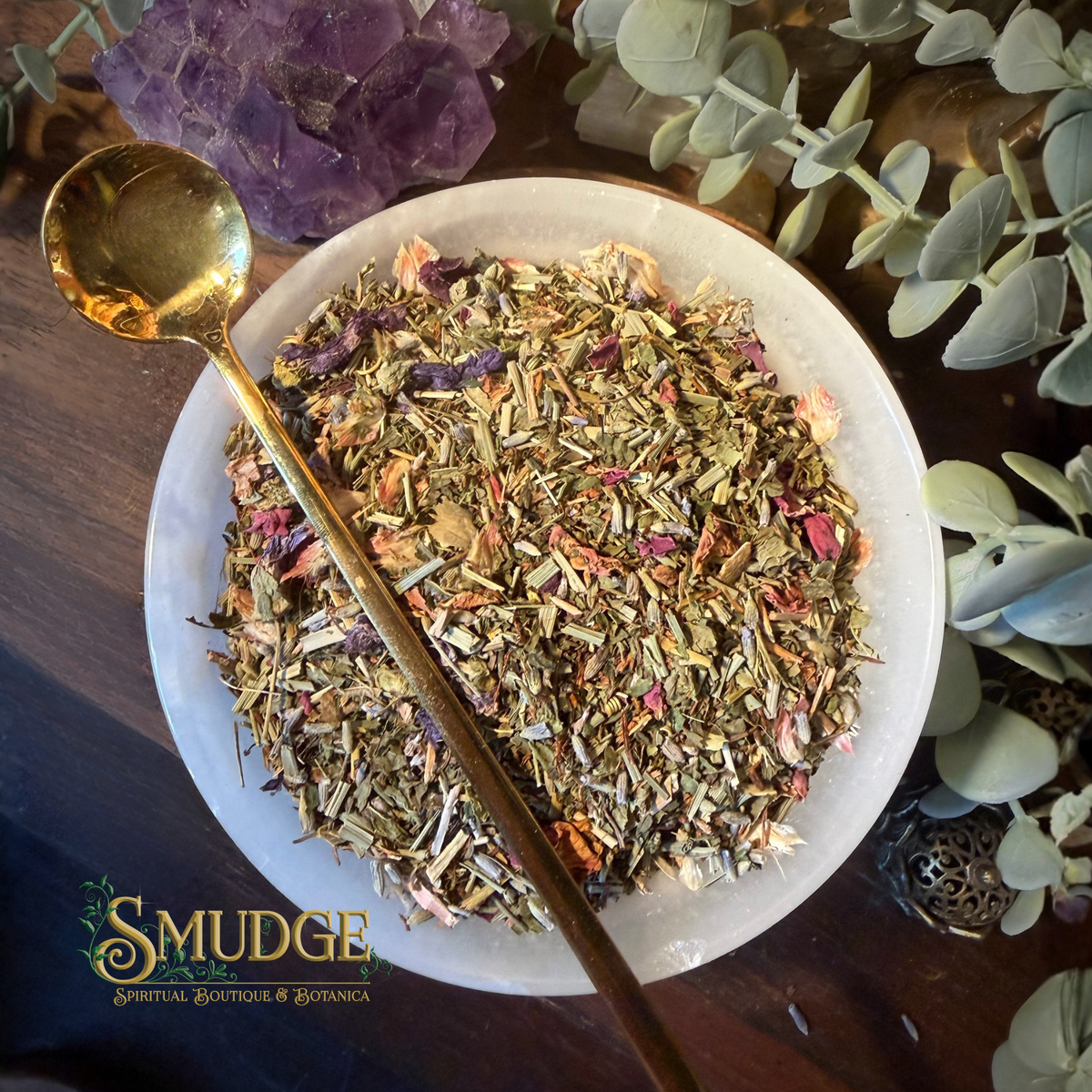 A top-down view of a white ceramic bowl filled with loose herbal tea or botanical blend, with a gold spoon resting across the bowl. The scene includes green leaves, purple crystals, and a dark wooden surface.