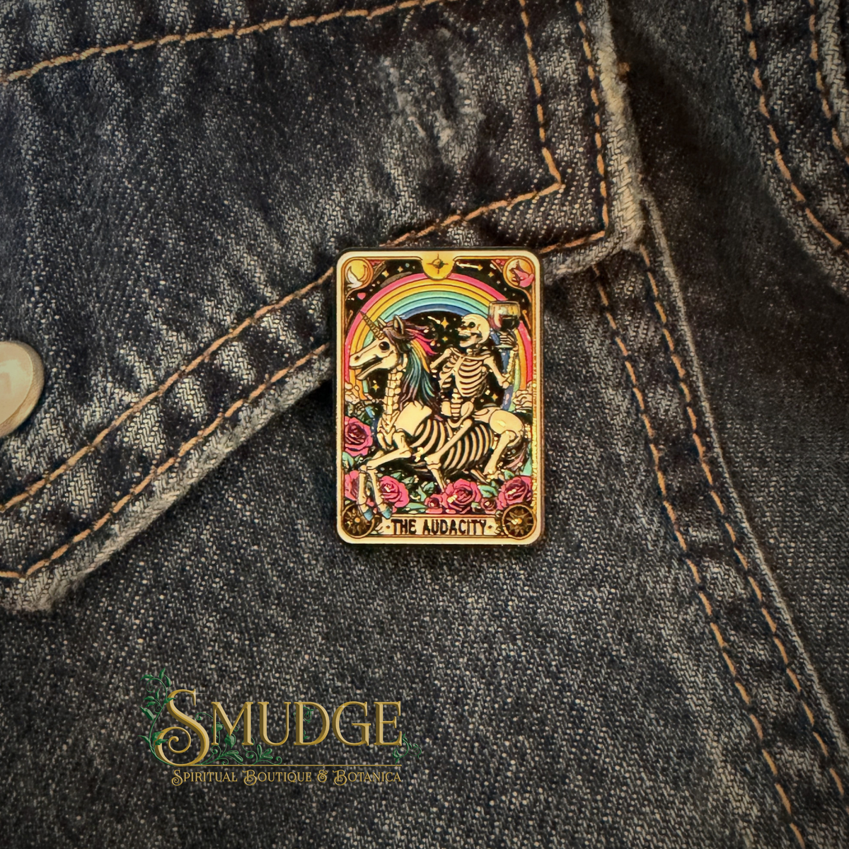 A rectangular enamel pin featuring a tarot-style skeleton rider under a rainbow and roses, pinned onto dark denim fabric. The pin has a gold-tone border and multicolored artwork.