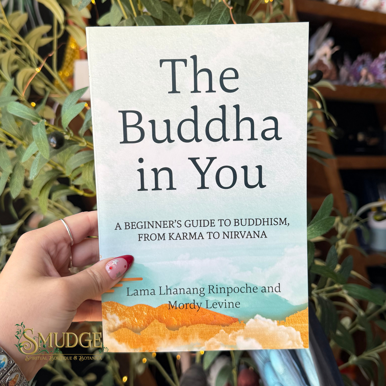 A hand holds a paperback book titled "The Buddha in You" in front of a background of green plants and shelves. The book cover has a light blue-gray watercolor texture with dark gray text and an orange and yellow mountain landscape illustration at the bottom.