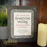 The Complete Shadow Work Workbook & Journal by Kelly Bramblett, displayed on a dark wooden surface next to a potted plant and a cream candle. The book features a gray and white mandala-style border with a white center containing black and red text.