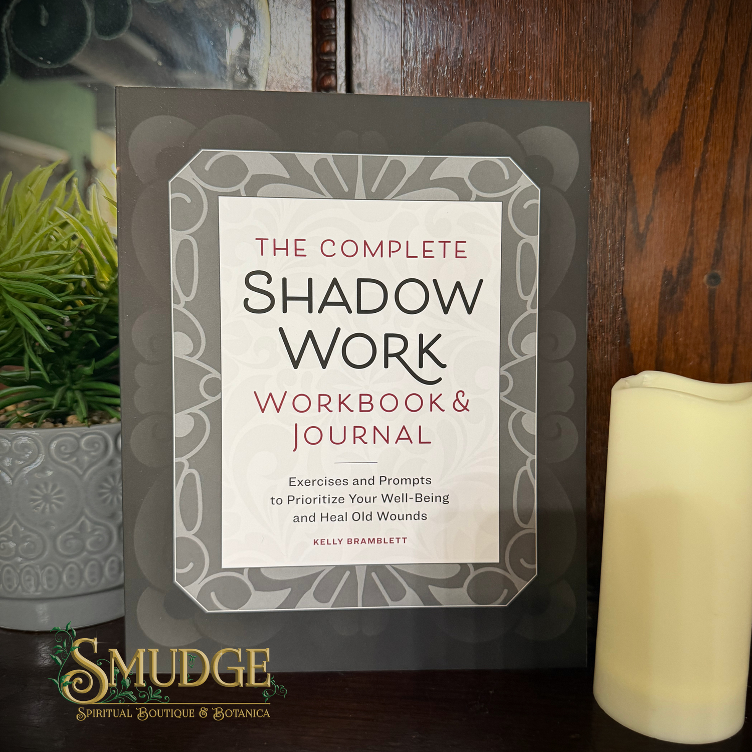 The Complete Shadow Work Workbook & Journal by Kelly Bramblett, displayed on a dark wooden surface next to a potted plant and a cream candle. The book features a gray and white mandala-style border with a white center containing black and red text.