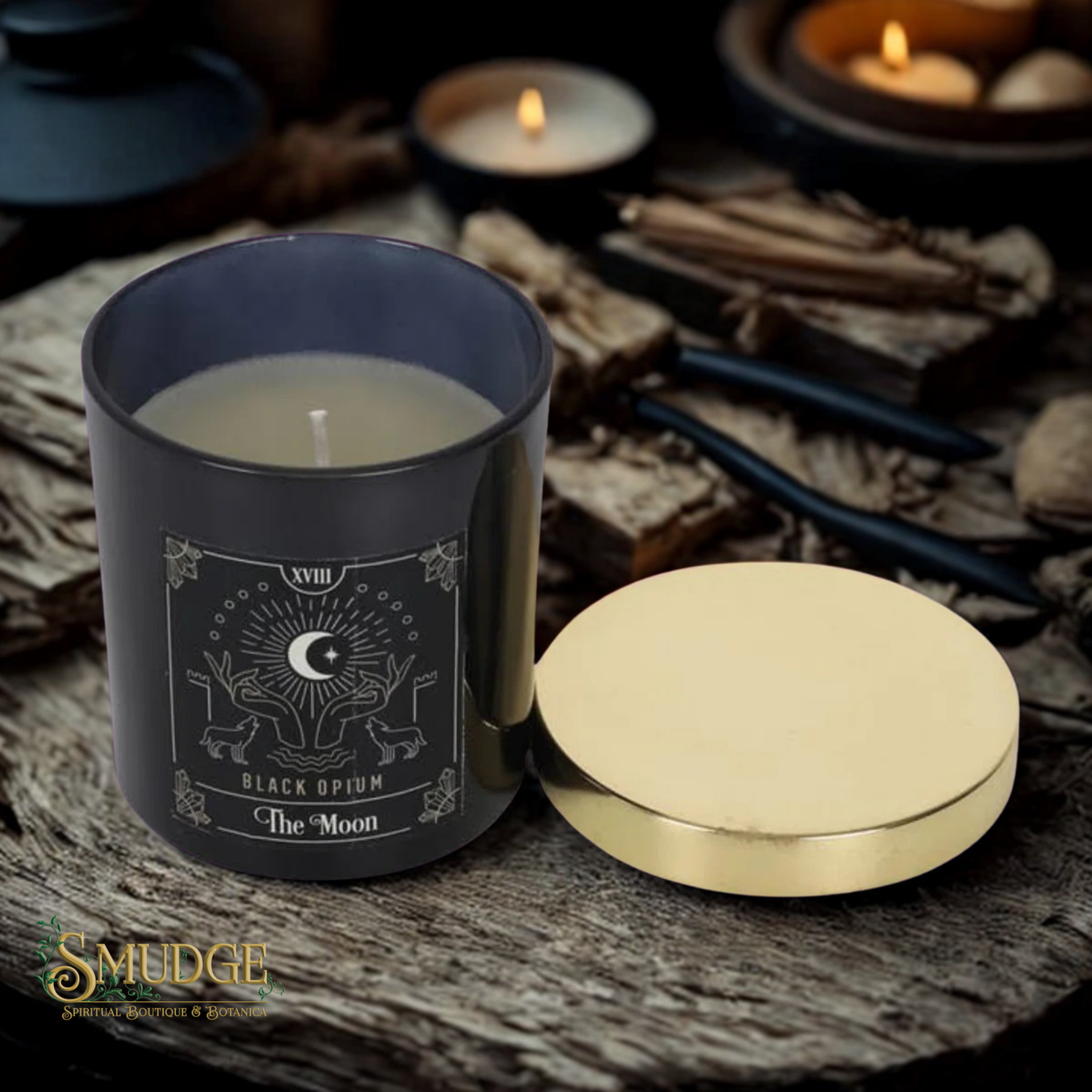 A dark gray glass candle jar labeled 'Black Opium' with 'The Moon' artwork, shown next to its gold-toned lid on a rustic wood surface with candles and dried herbs in the background.