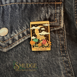 A rectangular enamel pin shaped like the 'The Moon' tarot card, featuring a gold-toned border, a crescent moon, clouds, stars, and colorful flowers, pinned onto a dark gray denim jacket. The brand name 'SMUDGE' is visible as a watermark in the lower-left corner.