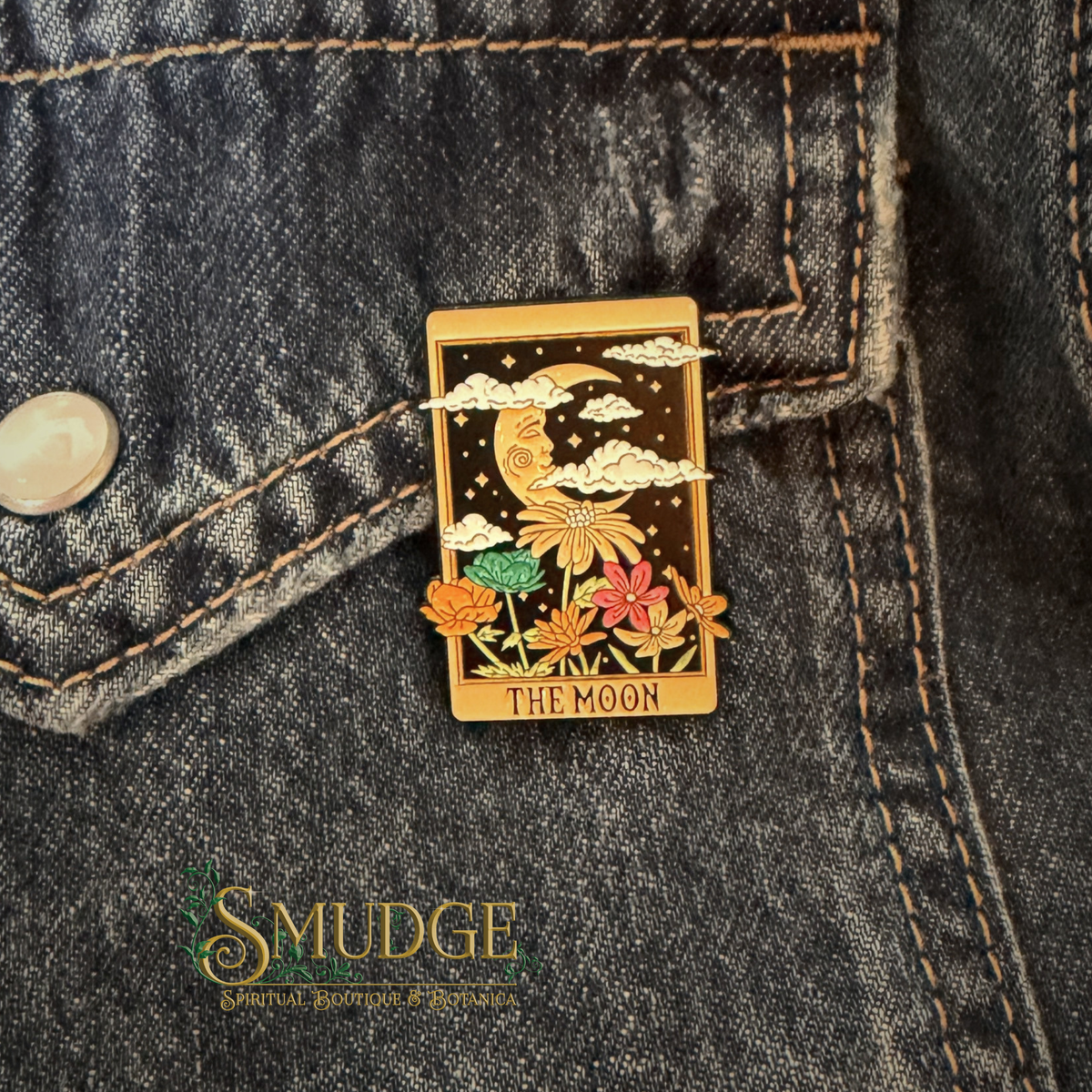 A rectangular enamel pin shaped like the 'The Moon' tarot card, featuring a gold-toned border, a crescent moon, clouds, stars, and colorful flowers, pinned onto a dark gray denim jacket. The brand name 'SMUDGE' is visible as a watermark in the lower-left corner.