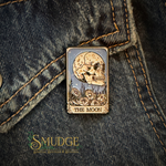 A rectangular enamel pin featuring a skull under a crescent moon with flowers at the bottom and the text 'THE MOON,' pinned onto dark blue denim fabric. The Smudge Handmade logo is visible in the lower-left corner.
