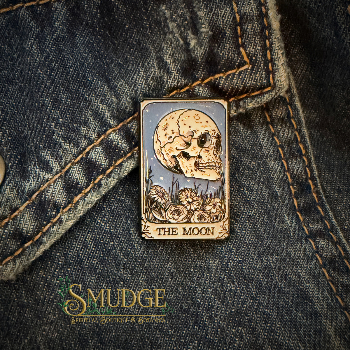 A rectangular enamel pin featuring a skull under a crescent moon with flowers at the bottom and the text 'THE MOON,' pinned onto dark blue denim fabric. The Smudge Handmade logo is visible in the lower-left corner.