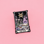 A tarot card enamel pin depicting 'The Star' card, featuring a gold star, an open book, and purple flowers on a black background, with a black ornate border, set against a solid pink background.