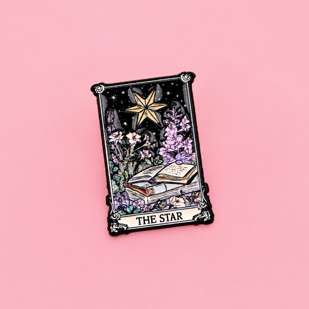 A tarot card enamel pin depicting 'The Star' card, featuring a gold star, an open book, and purple flowers on a black background, with a black ornate border, set against a solid pink background.