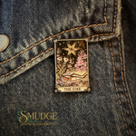 A rectangular enamel pin depicting 'The Star' tarot card, featuring a gold-toned star, open book, and pink flowers, pinned onto a dark gray denim jacket. The 'SMUDGE' brand logo is visible in the bottom-left corner.
