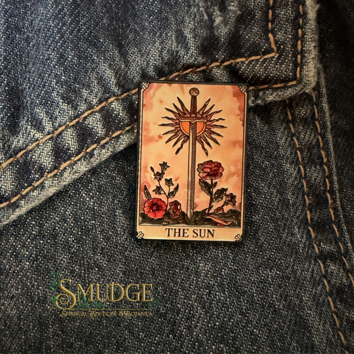A rectangular enamel pin depicting the 'The Sun' tarot card, pinned onto a dark blue denim jacket. The pin features a golden sun and sword with red flowers on a warm, aged beige background.