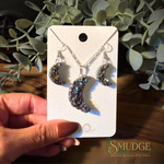 A hand holds a white display card featuring a silver necklace and matching earrings with crescent moon charms covered in iridescent, glittery resin. The background shows green eucalyptus leaves and a wooden surface, with the Smudge Spiritual Goods & Botanica logo in the bottom right corner.
