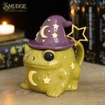 A whimsical wizard frog figurine in glossy olive green with a purple witch hat, gold crescent moon, and gold stars, set on a wooden surface with a blurred background of candles and books.