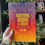 Book titled 'Traditional Hoodoo & Conjure' held by a hand with green nail polish, surrounded by plants.