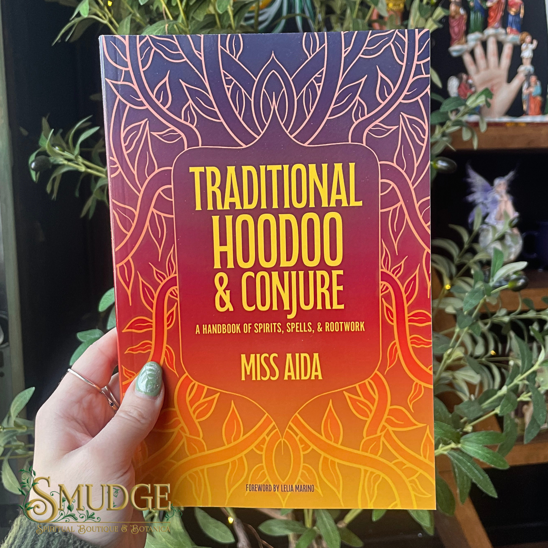 Book titled 'Traditional Hoodoo & Conjure' held by a hand with green nail polish, surrounded by plants.