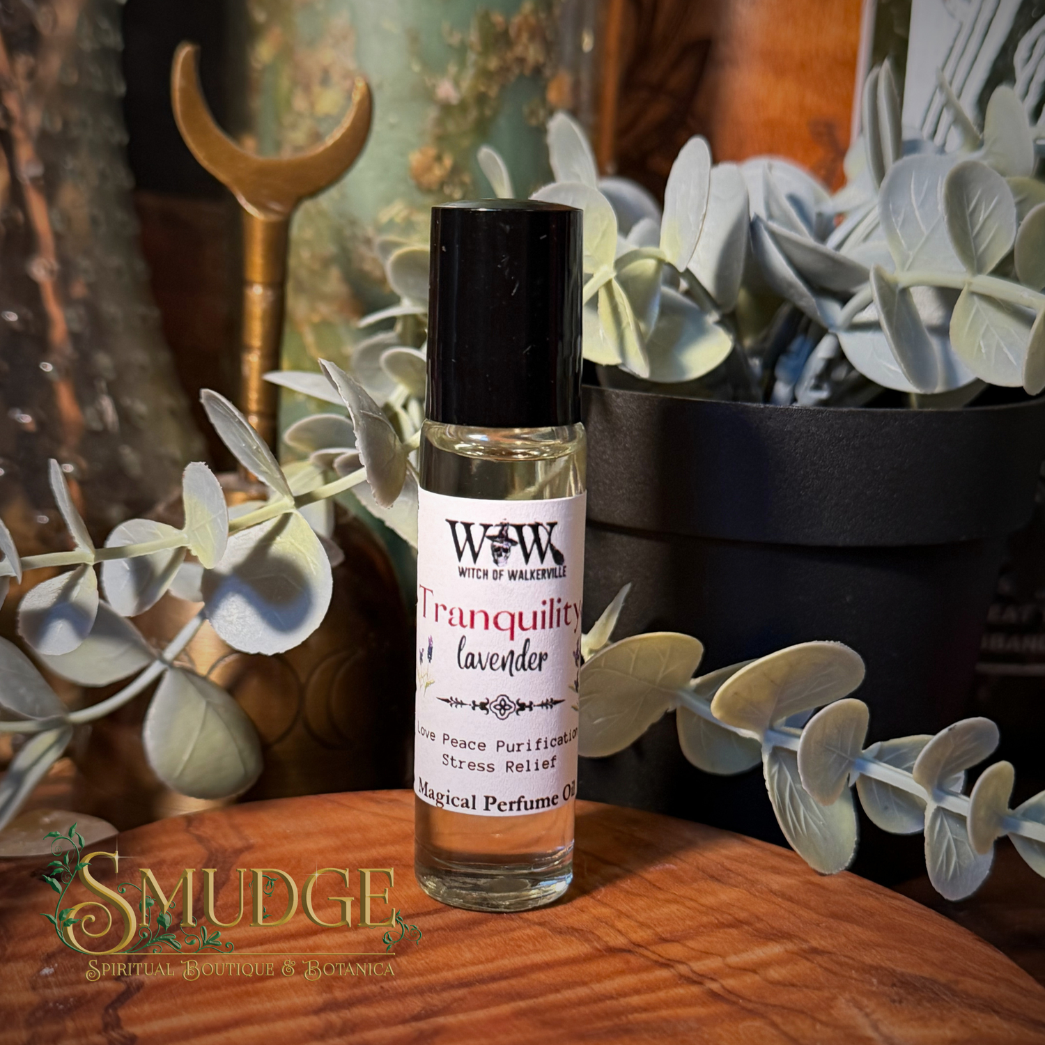 A small clear glass perfume roller bottle with a black cap and a white label reading 'Witch of Malachite Tranquility Lavender' sits on a wooden surface surrounded by eucalyptus leaves and decorative props.
