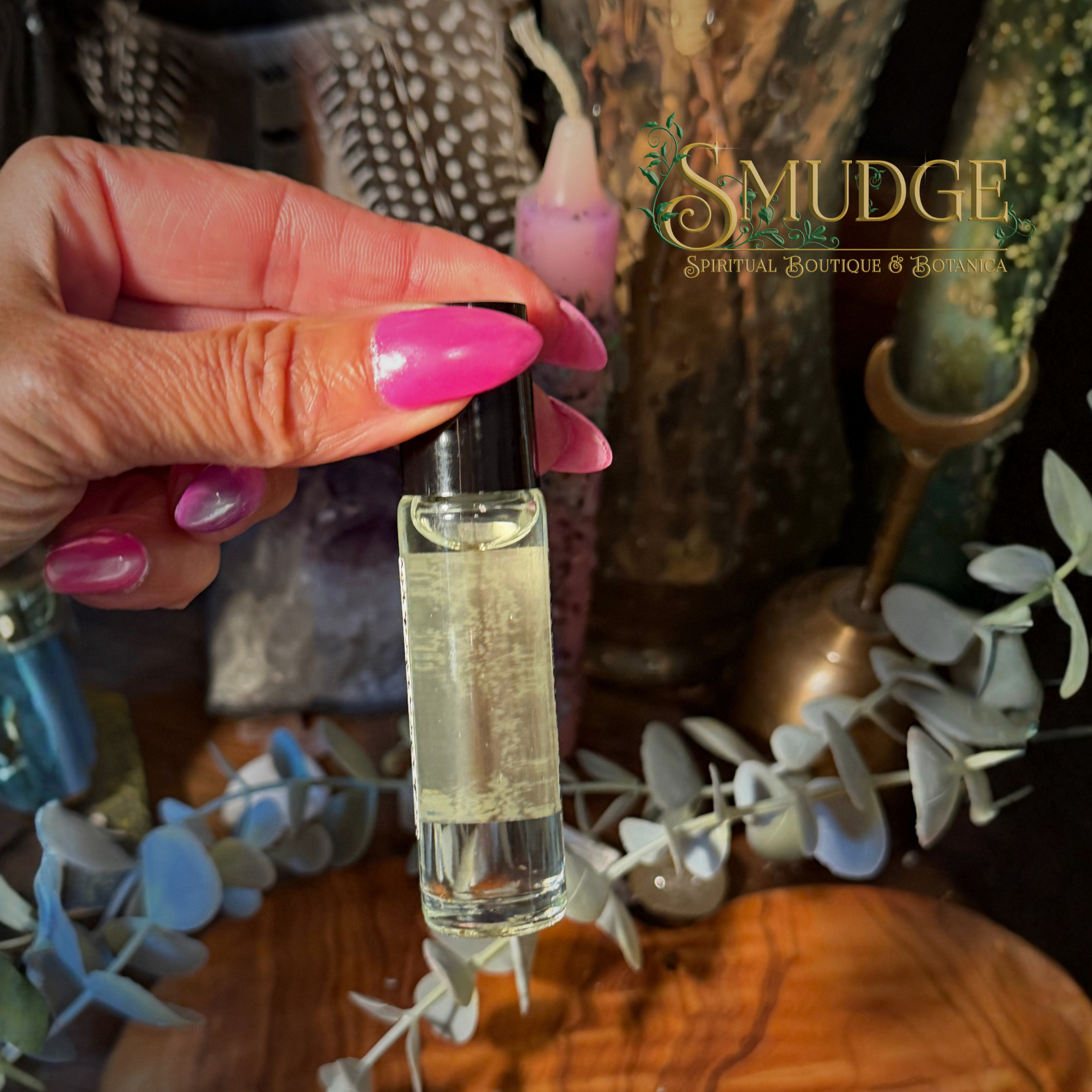A hand with pink nails holds a small clear glass roller bottle filled with pale yellow liquid. The background is a styled tabletop scene with dried flowers, greenery, and a visible 'SMUDGE Spiritual Boutique & Botanicals' logo watermark.